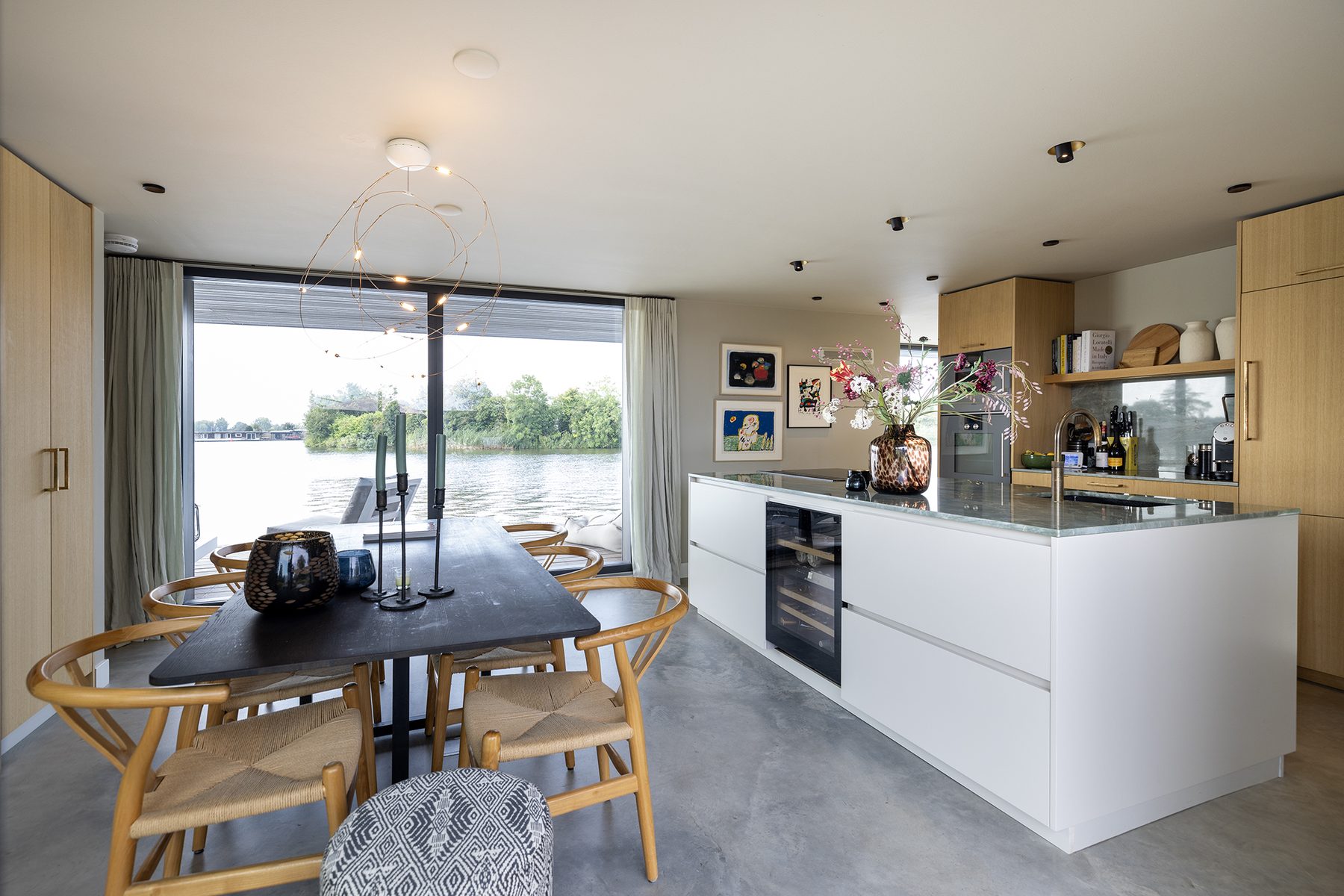 Eetkeuken van de woonark met kookeiland, eettafel en doorlopende glasgevel over het water